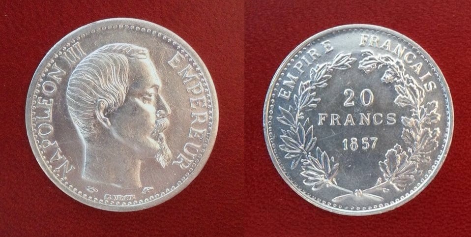 A photograph of a French 1857 20 Franc coin with the face of a man on one side and text saying 20 Francs on the other. The coin is made of aluminum and is shiny.