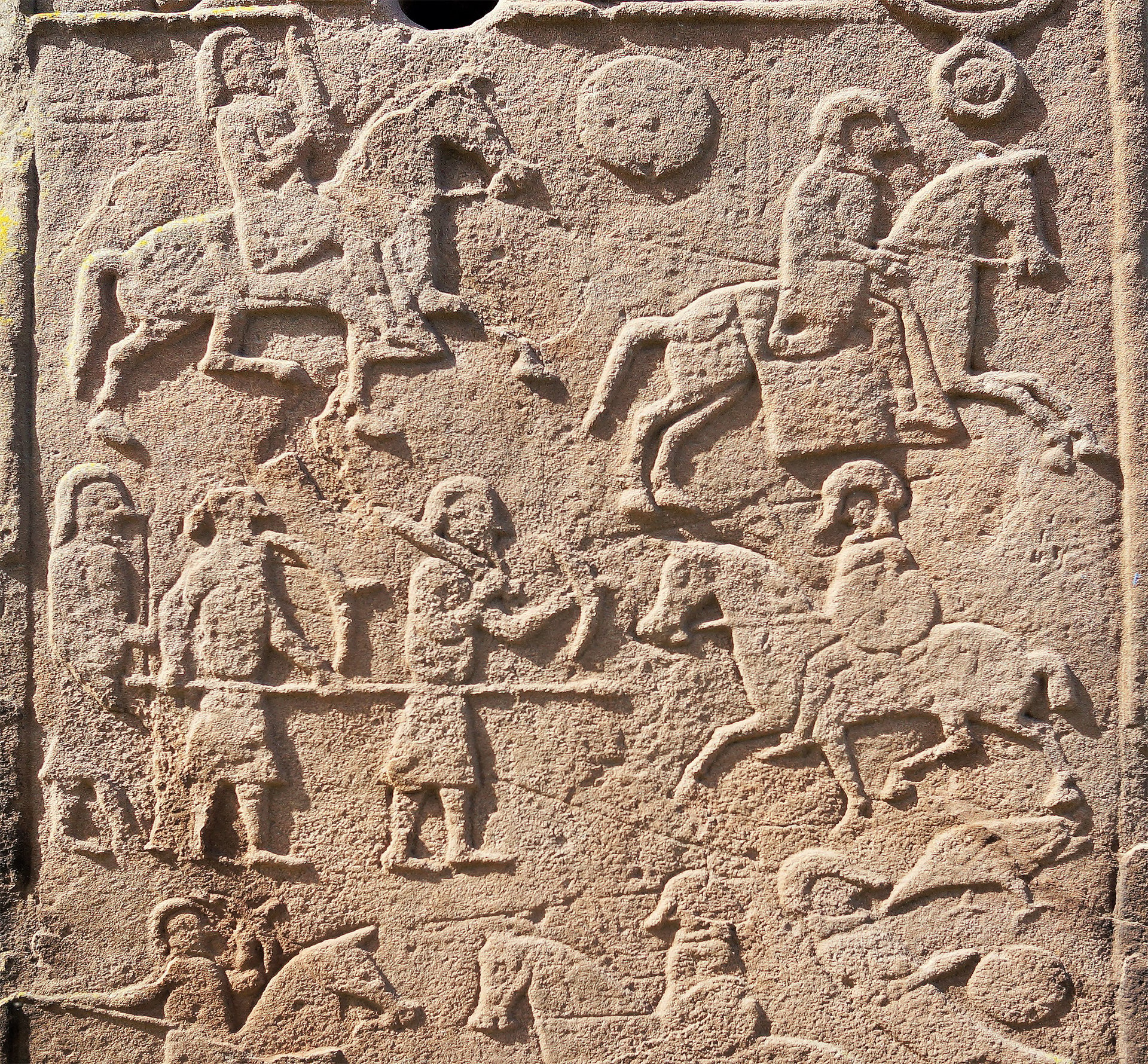 A photograph of a Pictish stone relief carving. The carving depicts a battle scene between the Picts and the Northumbrians in the 7th century. The carving shows horses and soldiers with helmets, shields, and spears.