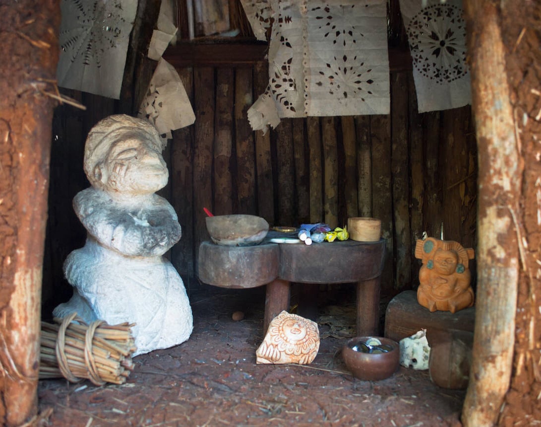A photo of a small wooden hut. Inside, there is a statue of a small smiling figure, an alux mythical creature, with folded arms, a small table with offerings, and a second statue of a figure with horns. The offerings on the table include a bowl, a candle, and a small blue bird figurine. The walls of the hut are made of wood logs and are decorated with paper cutouts.
