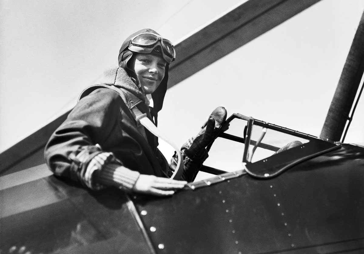 This is a black and white photograph of a woman, Amelia Earhart, in a pilot's uniform sitting in the cockpit of an airplane. She is wearing goggles and a helmet, and is smiling. The woman is holding onto the steering wheel with one hand and has his other arm resting on the side of the cockpit.