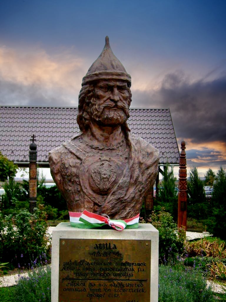 A photograph of a bust of Attila the Hun, a 5th-century ruler of the Hunnic Empire. The bust is made of bronze and shows Attila’s head and shoulders. He has long hair, a thick beard, and a stern expression. He is wearing a helmet with horns and a fur collar. The bust is on a stone pedestal with a plaque.