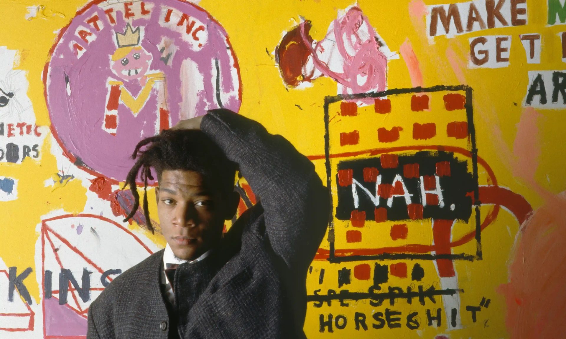 A photo showing artist Jean-Michel Basquiat standing in front of his vibrant artwork filled with bold strokes of yellow, red, black, and pink colors.