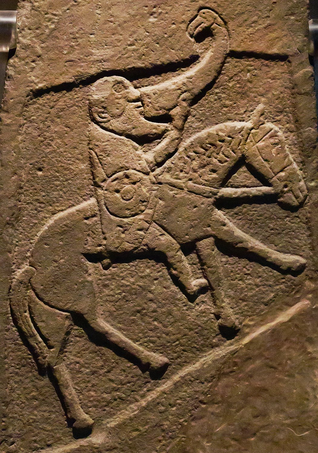 A carved stone slab showing a Pictish warrior on a horse. He has a bald head, a long beard. He drinks from a large horn that has a bird’s head at the end. The stone is dark gray and has some cracks.