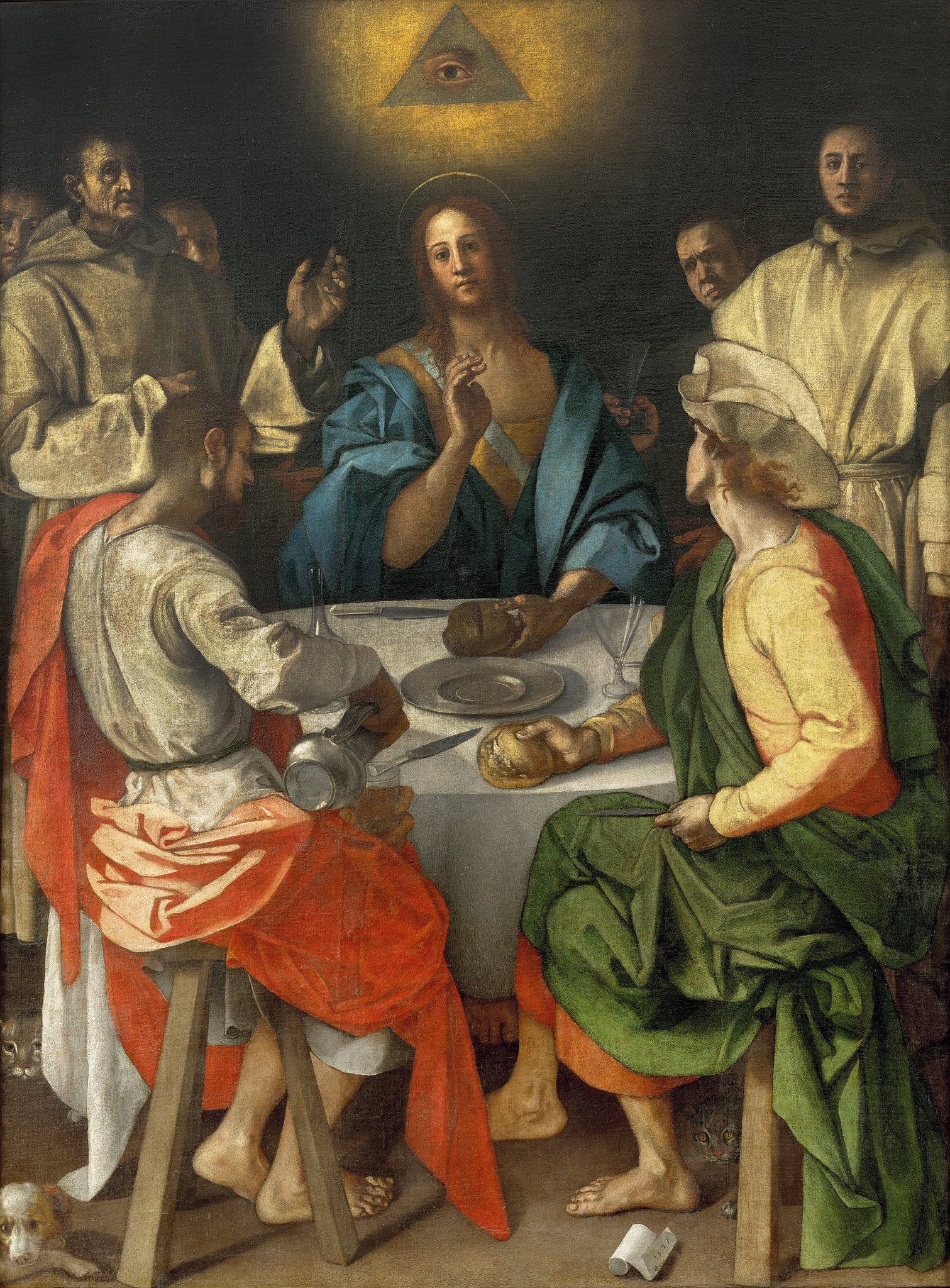 The painting shows a scene from the Bible, where Jesus reveals himself to two of his disciples after his resurrection. The painting is in a dark and somber tone, with a bright light coming from a window on the left. The light illuminates the face of Jesus, who is sitting at the center of a table, holding a piece of bread in his hands. He is wearing a blue robe and a red cloak, and has a halo around his head. He is looking at the viewer with a calm and serene expression. On his right, there is a man wearing a yellow robe and a green hat, who is leaning forward with his hands on the table. He is one of the disciples, named Cleopas, and he seems surprised and amazed by Jesus’ appearance. On his left, there is another man wearing a brown robe and a large hat with three points. He is the other disciple, named Luke, and he is looking at Jesus with curiosity and awe. Behind them, there are five other men who are watching the scene from a distance. They are monks from the Galluzzo Charterhouse, where the painting was commissioned. One of them, on the far left, is Leonardo Buonafede, the prior of the charterhouse, who is raising his left hand in a gesture that echoes Jesus’. The table has a white cloth on it, and some simple objects such as a plate, a bowl, a pitcher, and two glasses. There is also a scroll on the table, which has Pontormo’s signature and date on it. Under the table, there are two cats and a dog that are looking for food scraps. On the floor, in the bottom right corner of the painting, there is a skull that symbolizes death and mortality. At the top of the painting, there is an eye inside a triangle that represents God the Father and the Holy Trinity.