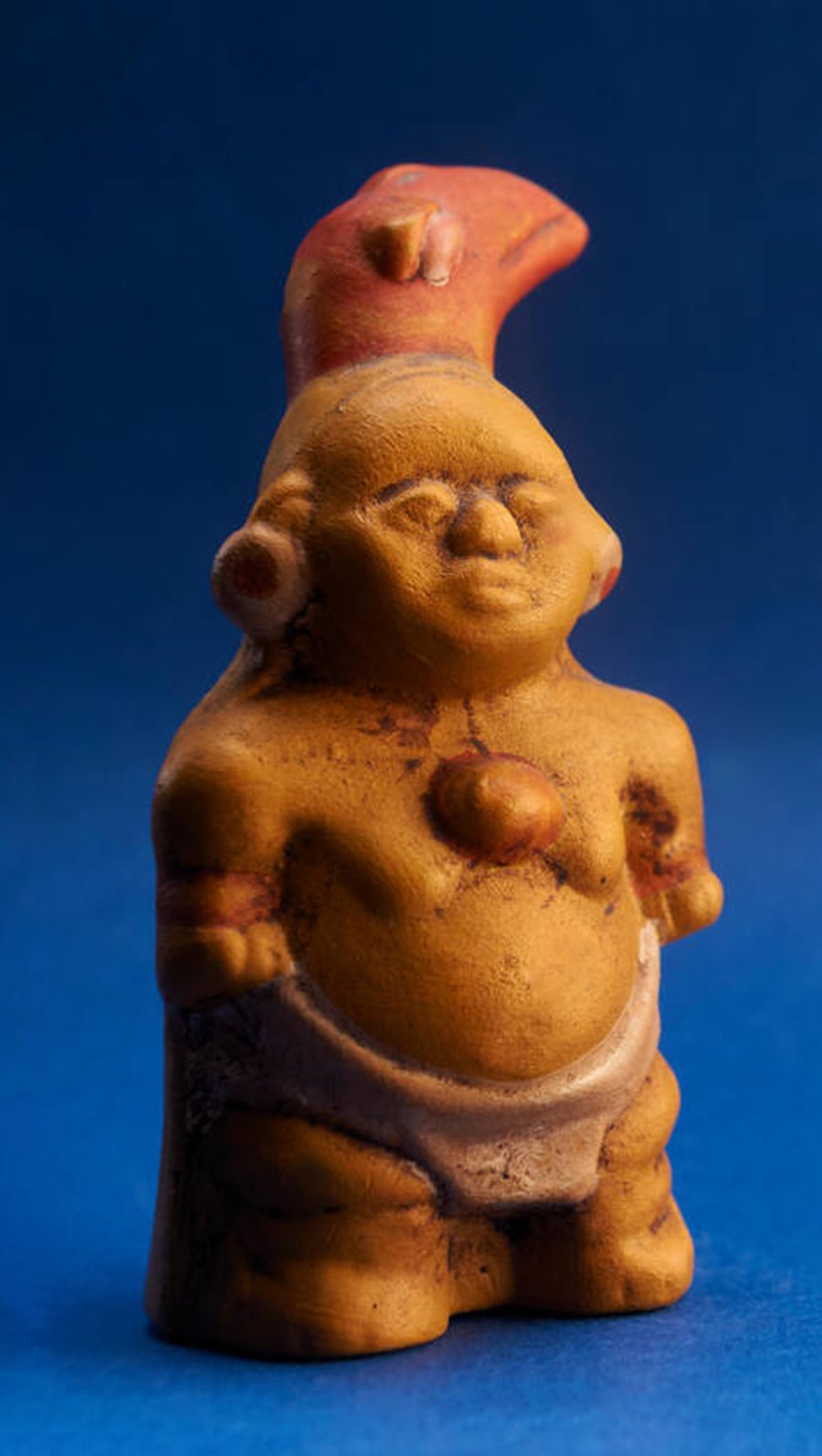 A photo of a small figurine depicting a chanque, a mythical little creature. The figurine is made of terracotta and has a round body and head. The chaneque is wearing a necklace and a loin cloth. The body of the figurine is covered in a brownish-yellow glaze. The background is a solid blue color.