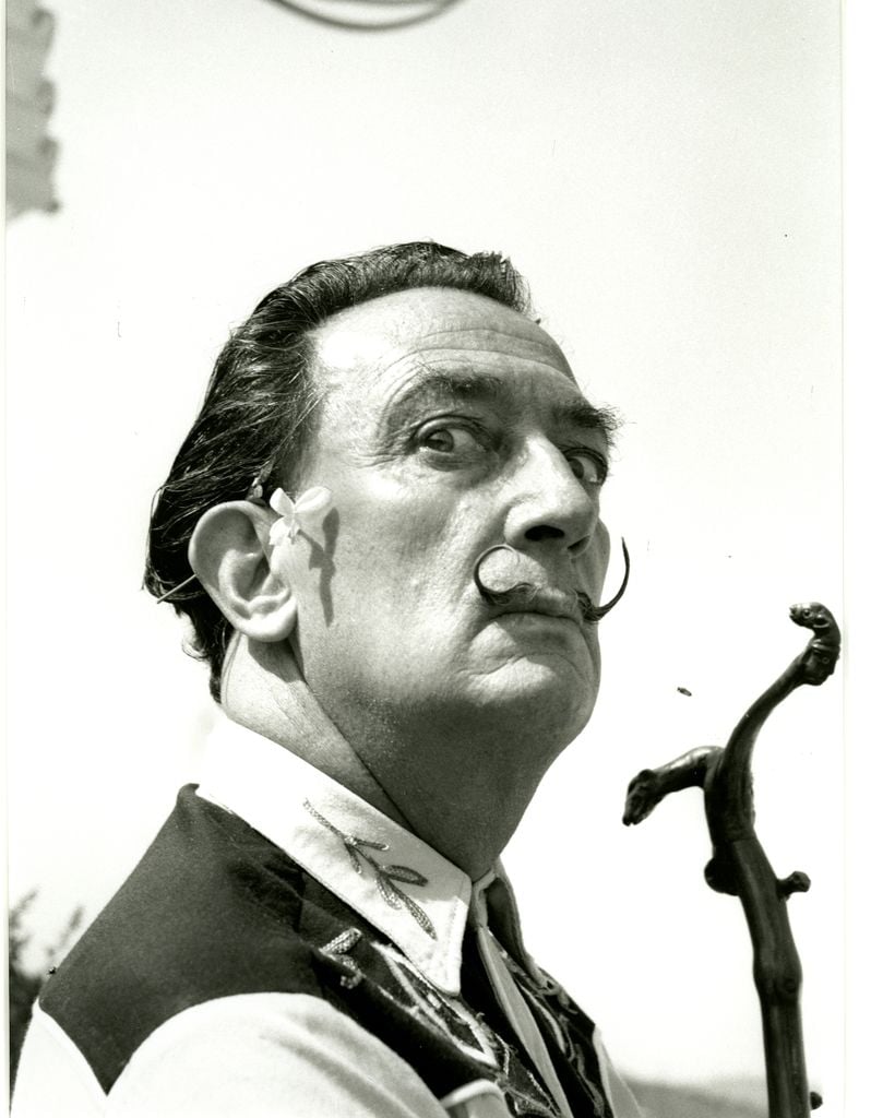 A black and white photo of a man's profile. The man has a long twirling mustache and a small flower behind his right ear. The man is holding a walking stick with a curved handle. The background is a clear sky. The man depicted is Salvador Dali, a famous Spanish surrealist painter and artist.
