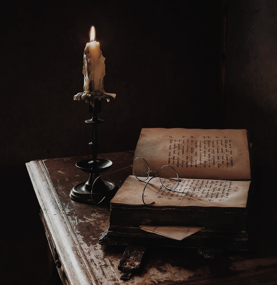 A photo of a night stand with a burning candle, open book and glasses in a dark room.