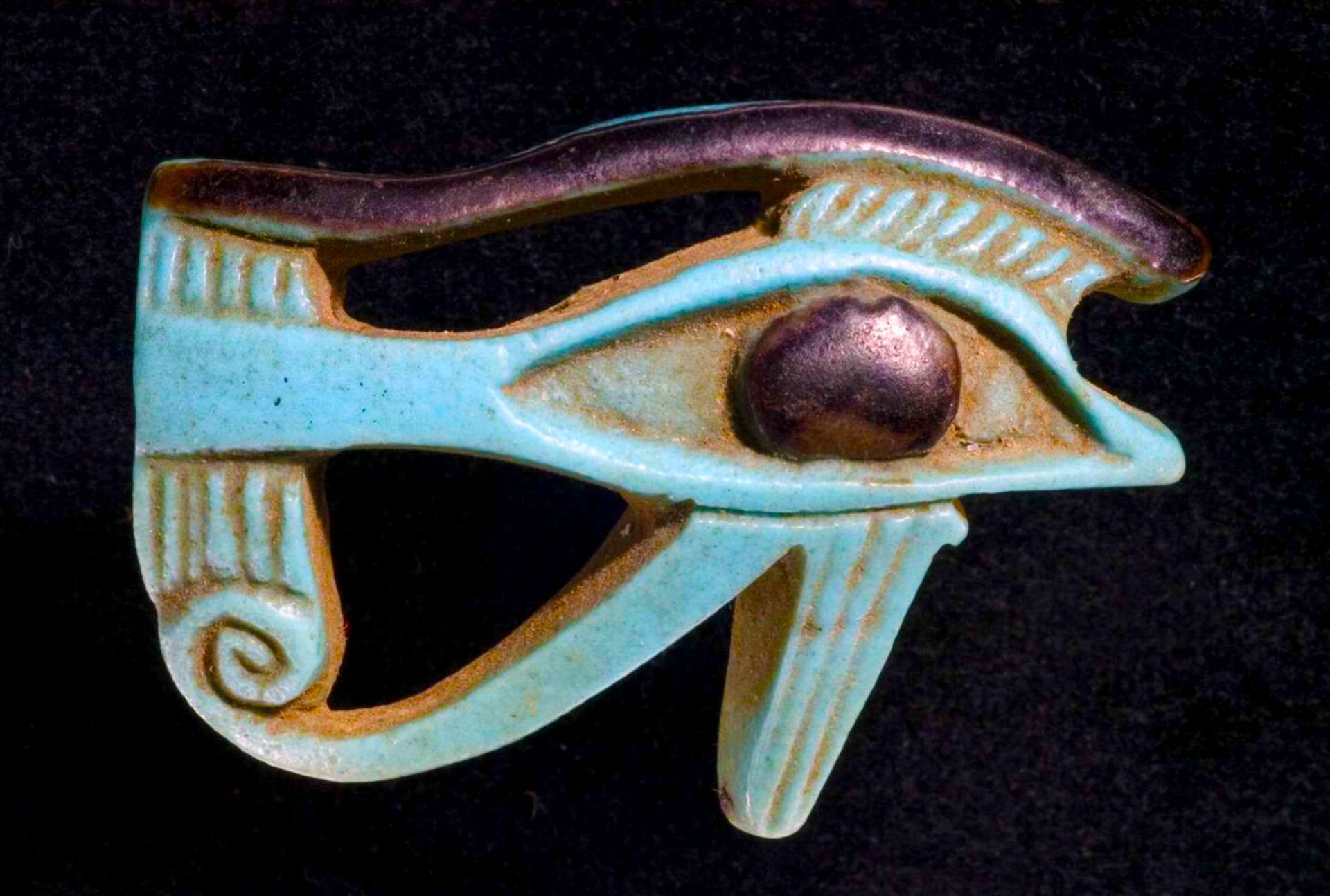 An ancient Egyptian Eye of Horus amulet made of turquoise faience with a brown pupil and a purple eyebrow on a black background.