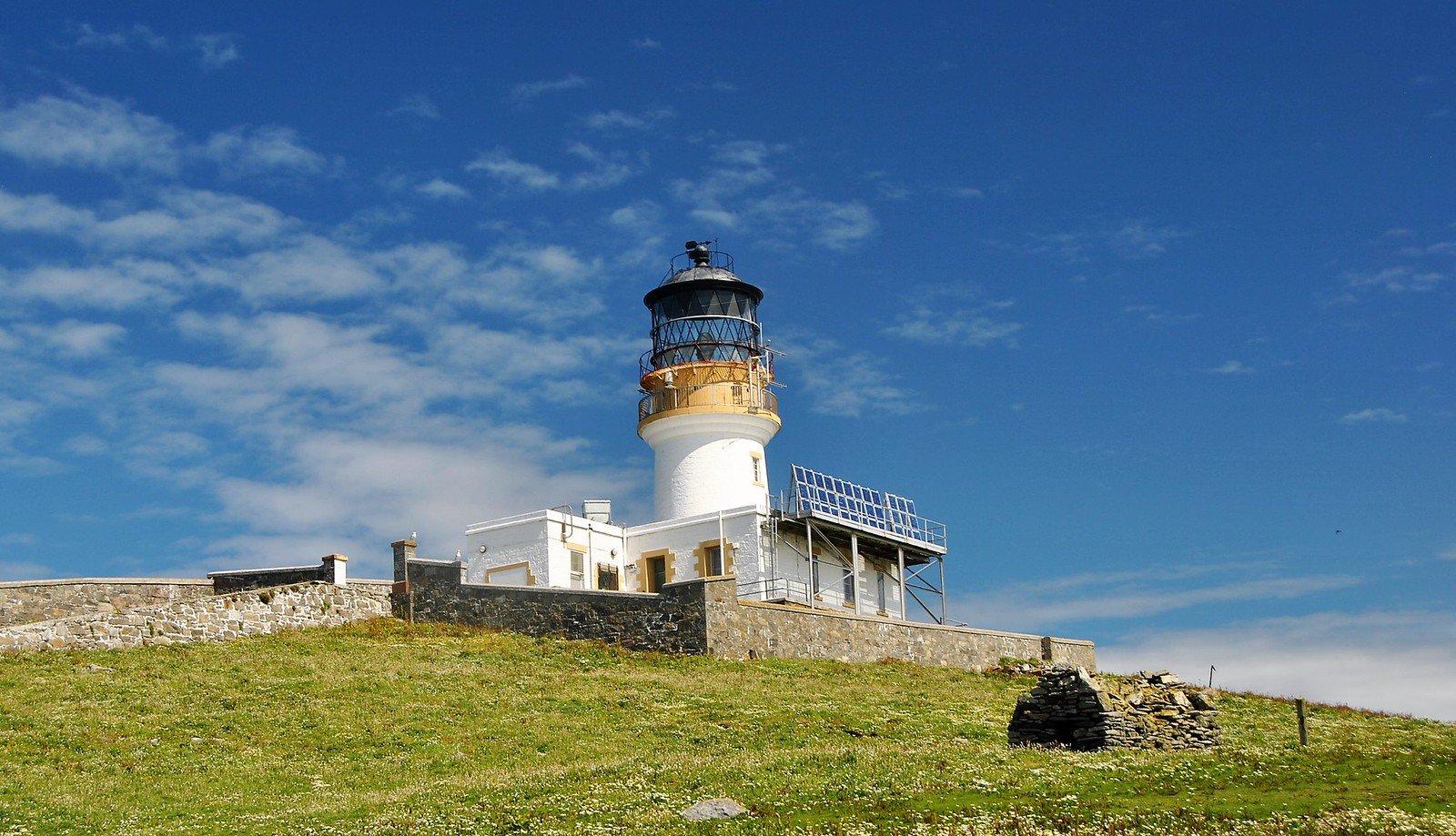 A photo realistic image of a white lighthouse with a black top and a solar panel on a grassy hill under a blue sky.
