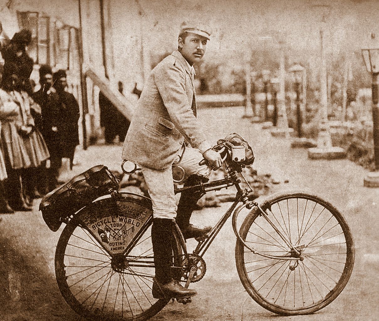 The black and white image shows a man in a suit and hat riding a bicycle. He is holding onto the handlebars with one hand. There are other people in the background.