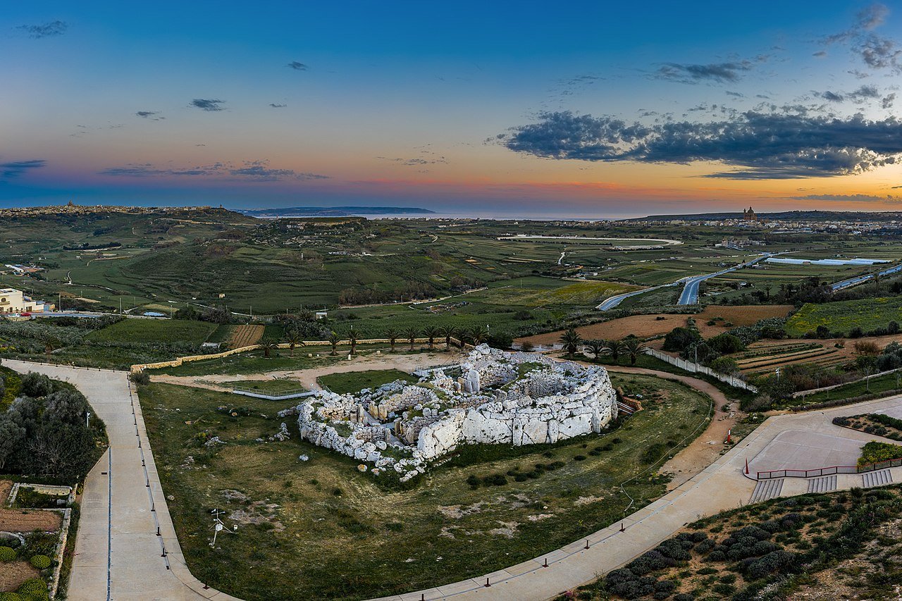 The image shows a beautiful view of ruins on a hill surrounded by green fields and a winding road. The ruins are made of white stone. The sky is orange and pink at sunset, casting a warm glow over the scene.