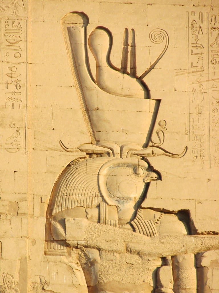 The image shows an ancient Egyptian hieroglyphic carving on a stone wall. The hieroglyphic is of the god Horus, a man with a falcon head. Horus is wearing a tall crown. The stone wall is weathered.