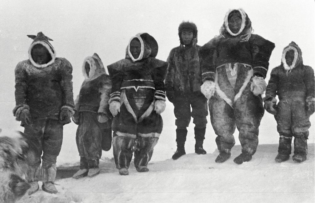 A group of Inuit people in traditional winter clothing standing on a snowy landscape with a dog.