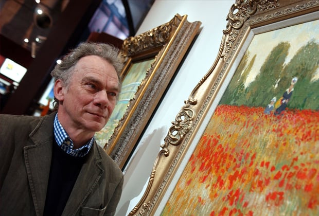 A photograph of a man standing in front of a painting of a field of poppies. He is wearing a blazer and sweater and has a lighthearted expression on his face. The painting is hanging on the wall behind him and there are other paintings visible in the background. The room is well lit. The room appears to be an art gallery.