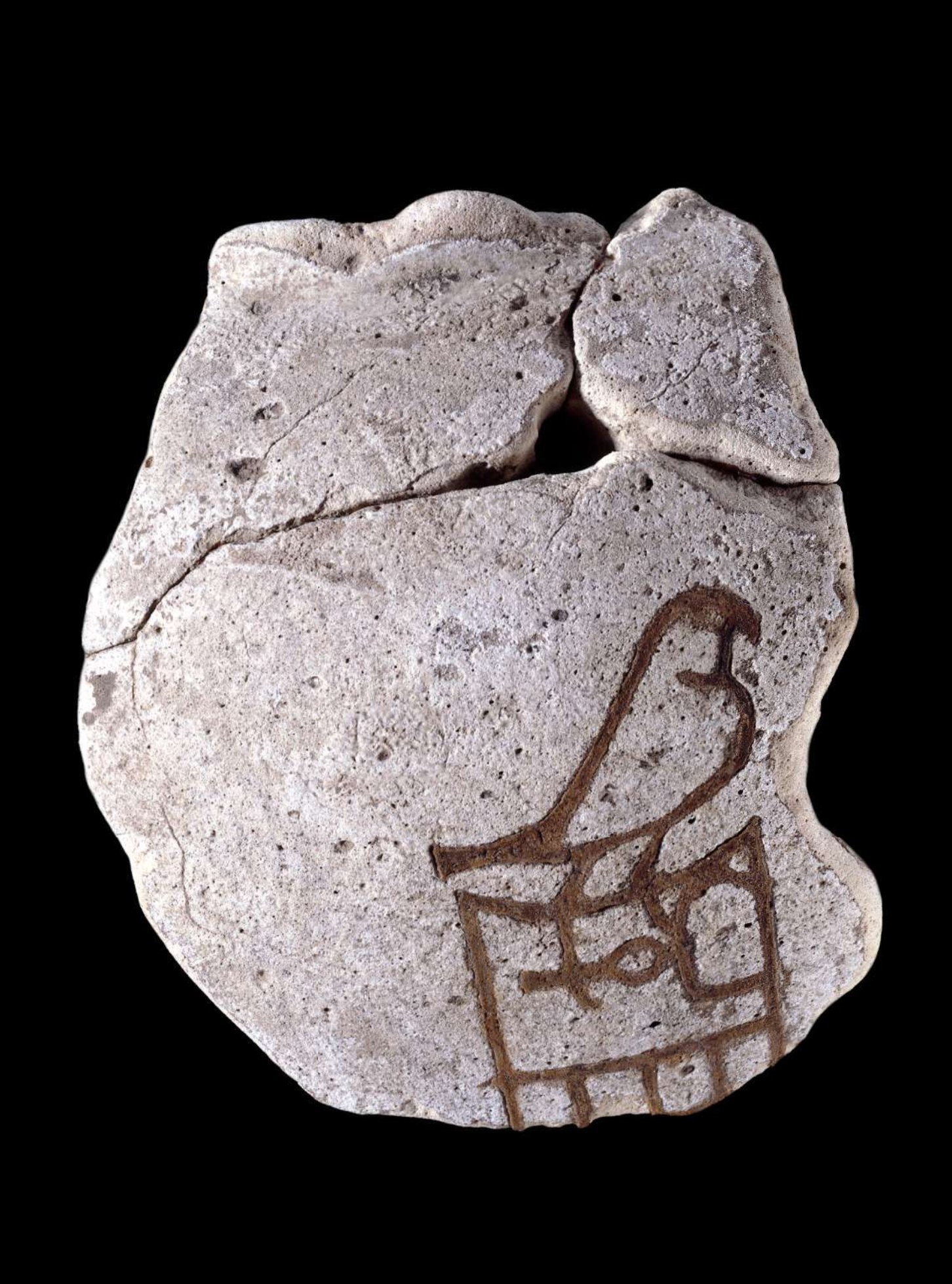 The image shows an ancient artifact that is a small piece of pottery. The pottery is a light beige color with a darker brown design on it. The design is of a falcon standing on a platform. The pottery is cracked and chipped in several places, indicating its age and fragility. The background is black, making the pottery stand out.