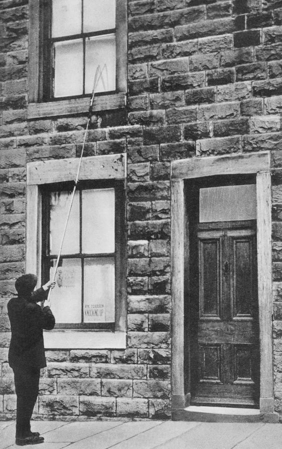 This black and white photograph shows a man standing in front of a brick building with a window on the second floor. The man is wearing a suit and holding a long rod in his hand. He appears to be a second floor window using the rod.