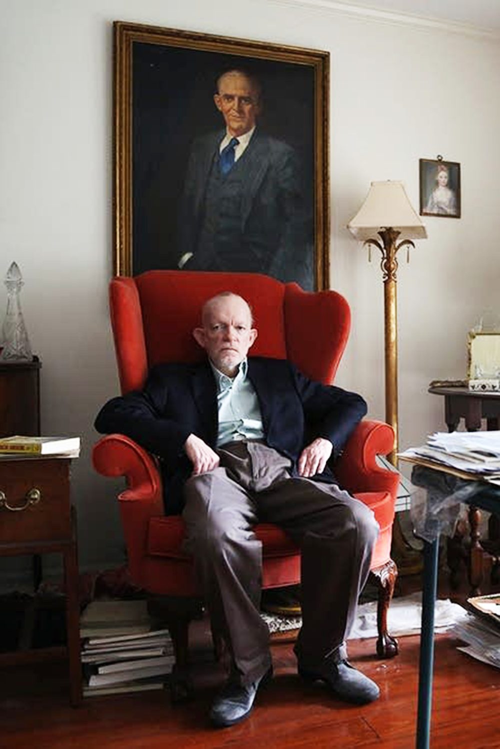 This photograph shows an older man sitting in a red armchair in a room with a large painting on the wall behind him. He is wearing a black suit and has a serious expression on his face. The room is cluttered with books and documents.