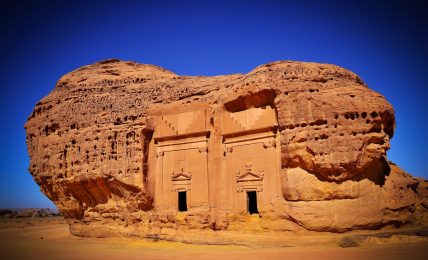 The image shows a large rock formation in the desert with two doors carved into it. The rock formation is surrounded by sand and there is a clear blue sky in the background.