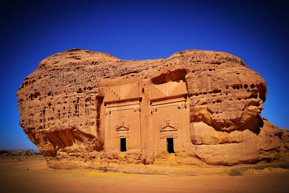 The image shows a large rock formation in the desert with two doors carved into it. The rock formation is surrounded by sand and there is a clear blue sky in the background.
