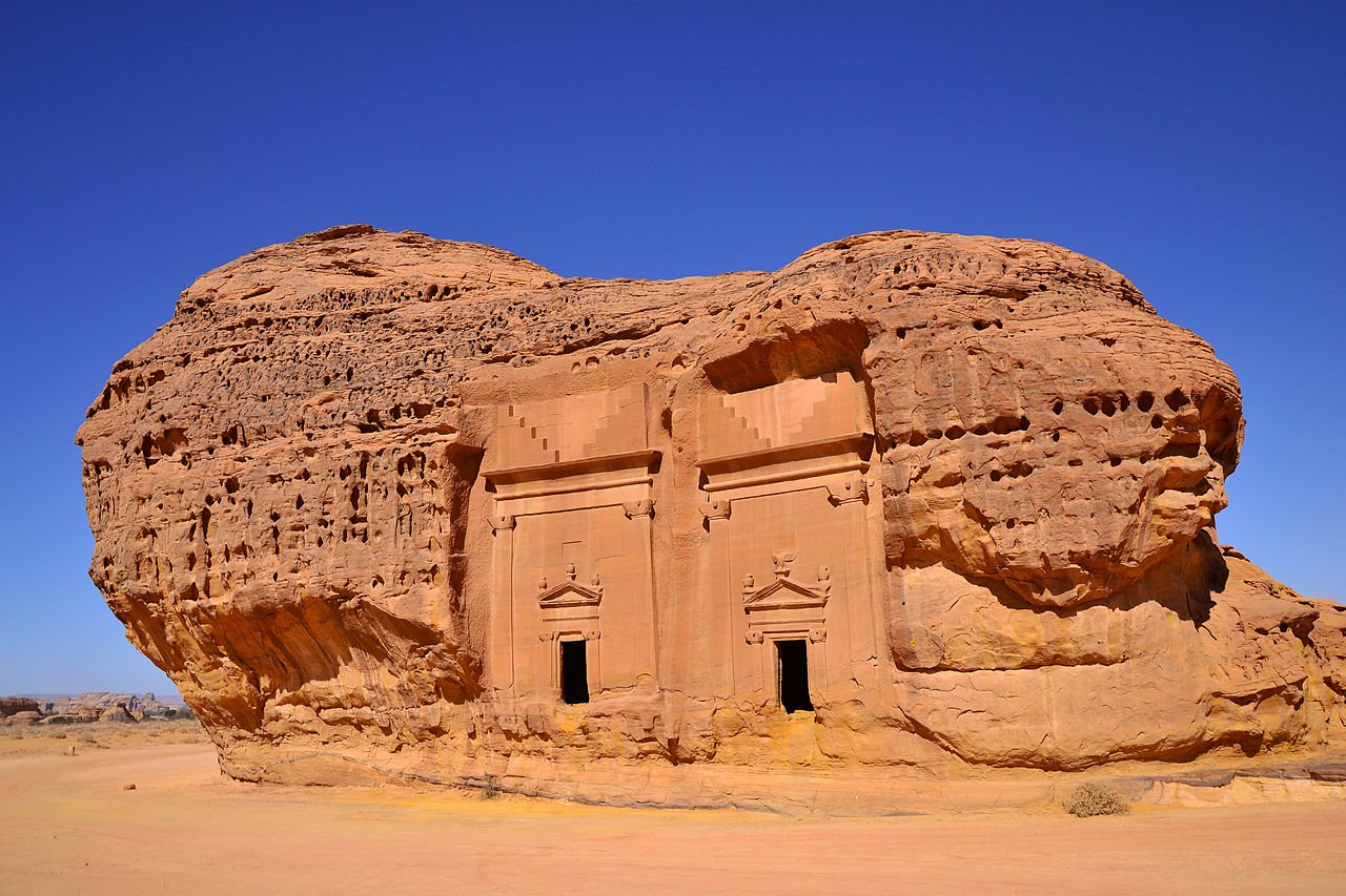 A photograph of a large rock formation in the desert, with a pair of doors carved into it. The rock formation is surrounded by sand and there is a clear blue sky in the background.