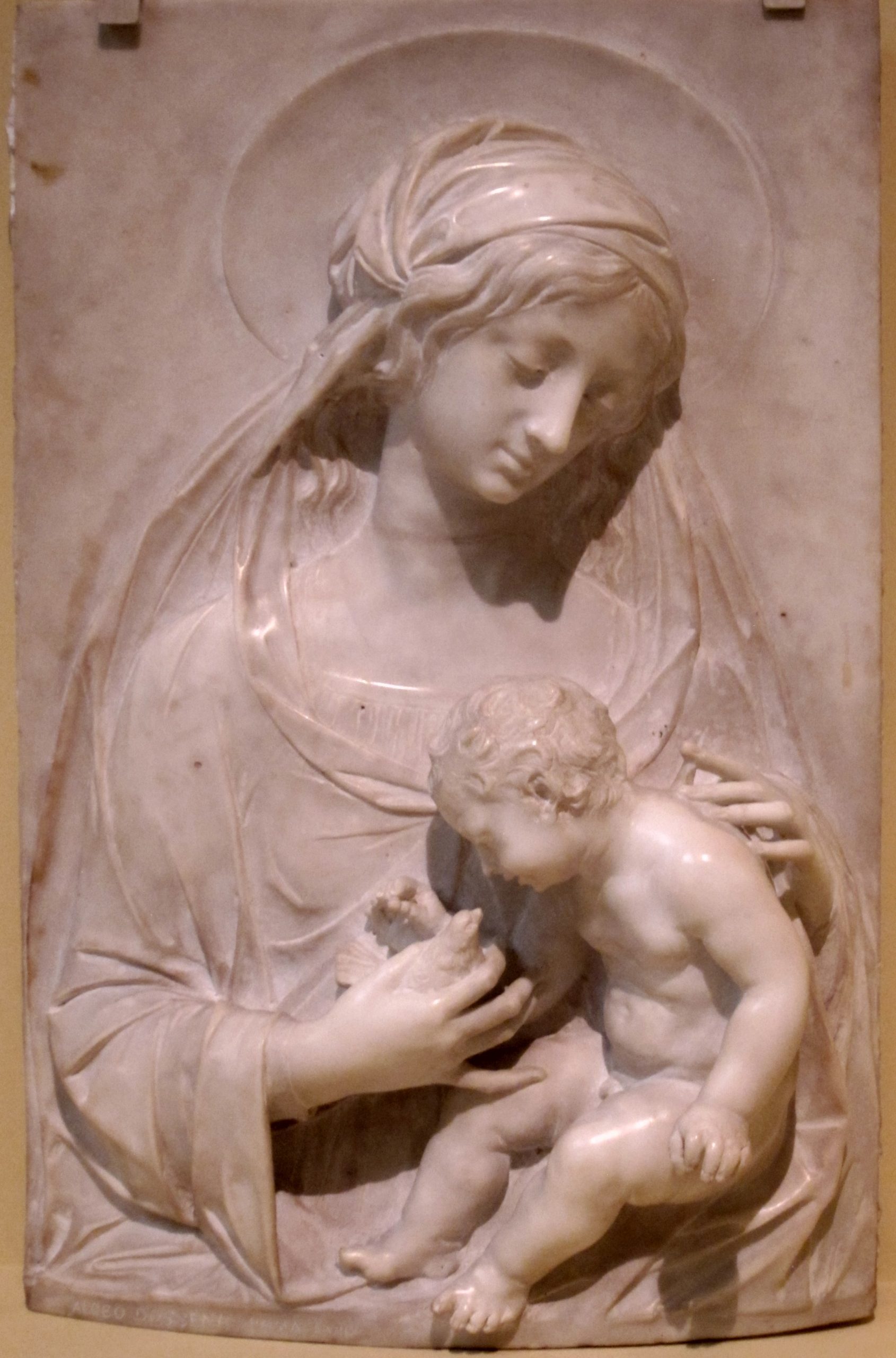 The image is a marble relief depicting the Virgin Mary holding a baby in her lap. The Virgin Mary is depicted with a serene expression and is dressed in a long flowing robe. The baby in her lap is depicted as a small, naked infant. The background of the relief is blank, with no other elements visible.