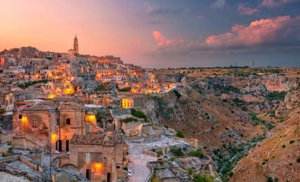 A high dynamic range, arial photograph of Matera, Italy at dusk. The sun is setting and the ancient city's lights are coming on.