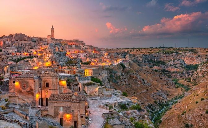 A high dynamic range, arial photograph of Matera, Italy at dusk. The sun is setting and the ancient city's lights are coming on.