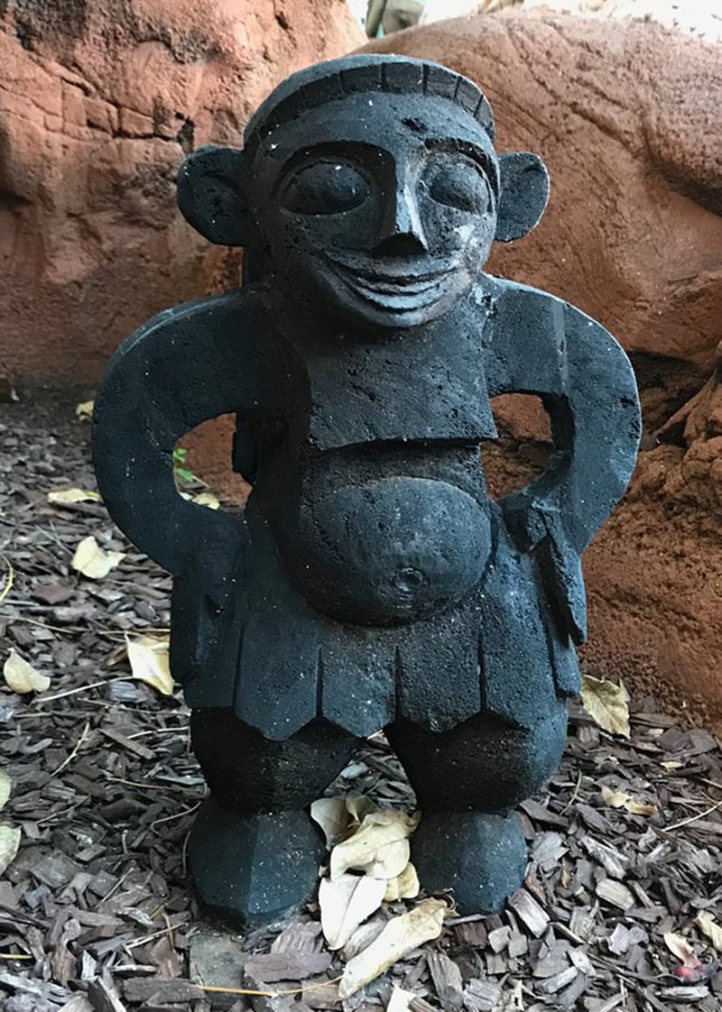 A photo of a black statue of a figure with a round head and a large belly. The figure is standing on a bed of brown mulch. The figure has a large round head with a smiling face and large round eyes. The figure has a large round belly and is wearing a skirt with triangular patterns. The background consists of a red rock wall.