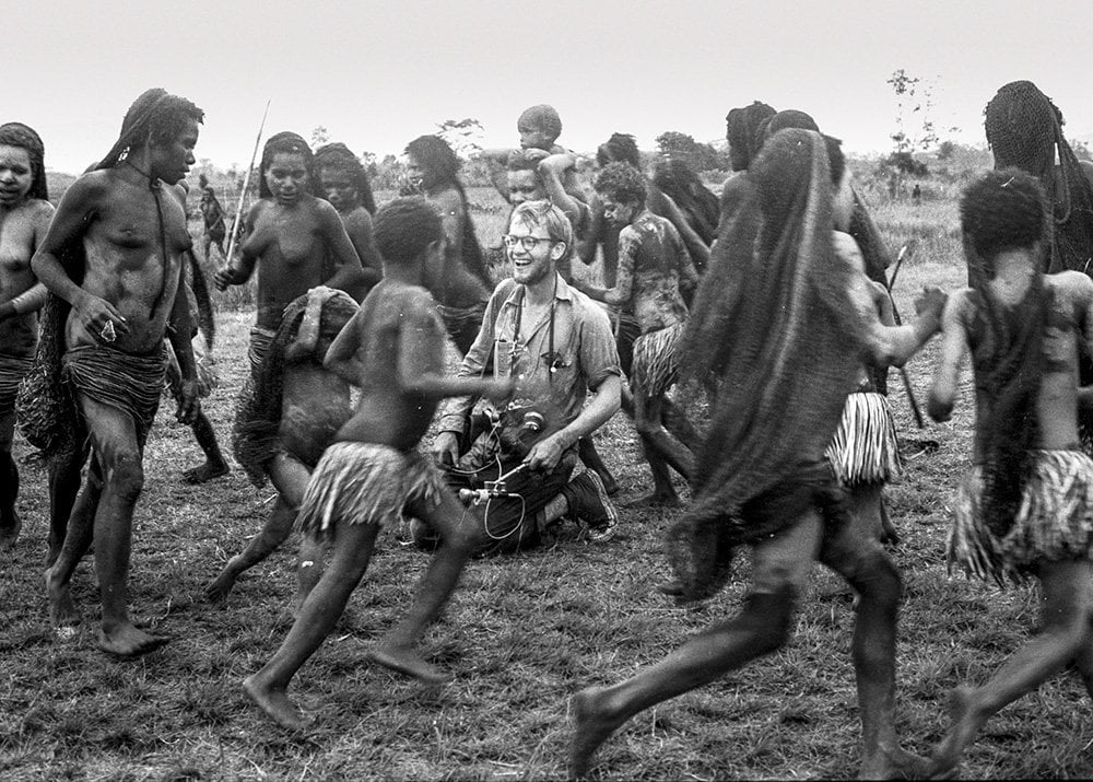 The image shows a group of people dressed in traditional African clothing, dancing and singing in a circle. They are all smiling and appear to be enjoying themselves. There is a man kneeling in the center of the group. The people in the group are all wearing head wraps and some have beaded jewelry around their necks. The background is a grassy field with trees in the distance.