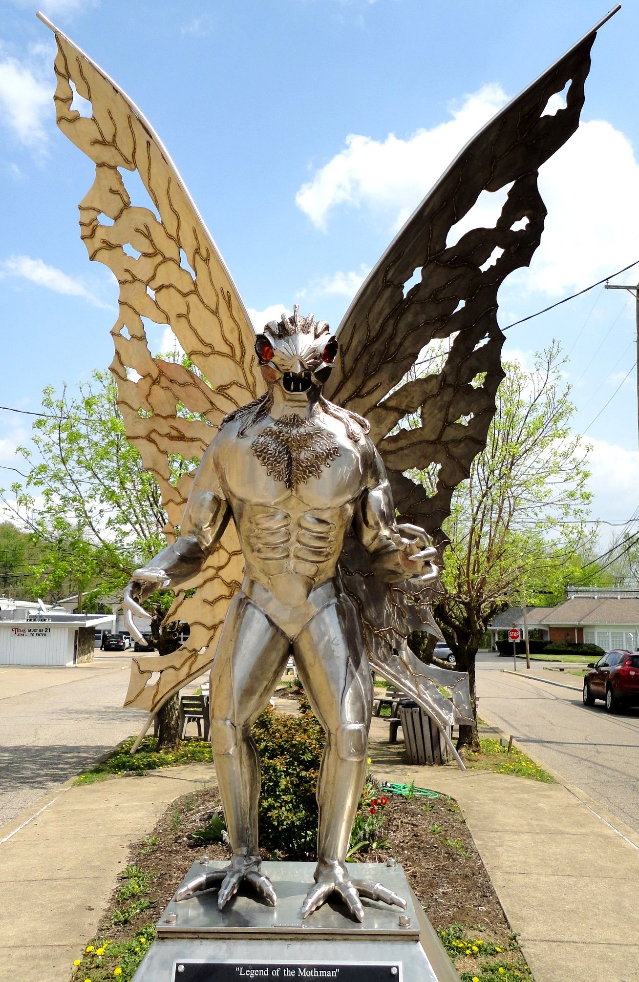 A photo of a metal statue of a winged creature with a humanoid body and a dragon-like head, standing on a pedestal with a plaque that reads "Legend of the Mothman". The background is a street with trees and buildings and a blue sky with clouds.