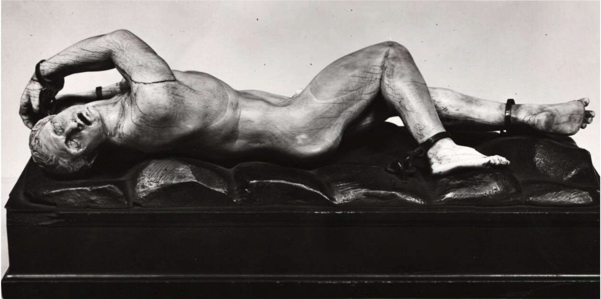 A black and white photo of a stone sculpture of a nude man lying on a black rock base.