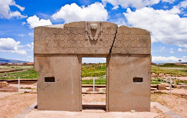 A photograph of a large stone doorway with a carving of a lion's head on the front. The structure appears to be in a desert setting with mountains in the background. The sky is clear and blue.