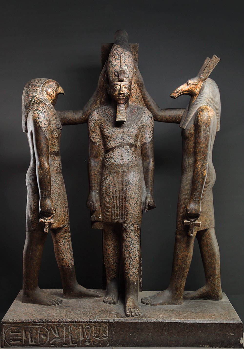 The image shows a statue of an ancient Egyptian pharaoh, Ramesses III, with two deities on either side. The pharaoh is standing in the center. The deities on either side of the pharaoh are Horus and Set. Horus is on the left side of the pharaoh and is depicted as a falcon-headed man. Set is on the right side of the pharaoh and is depicted as a jackal-headed man. The statue is made of stone and is in a dark gray color. The background is black.