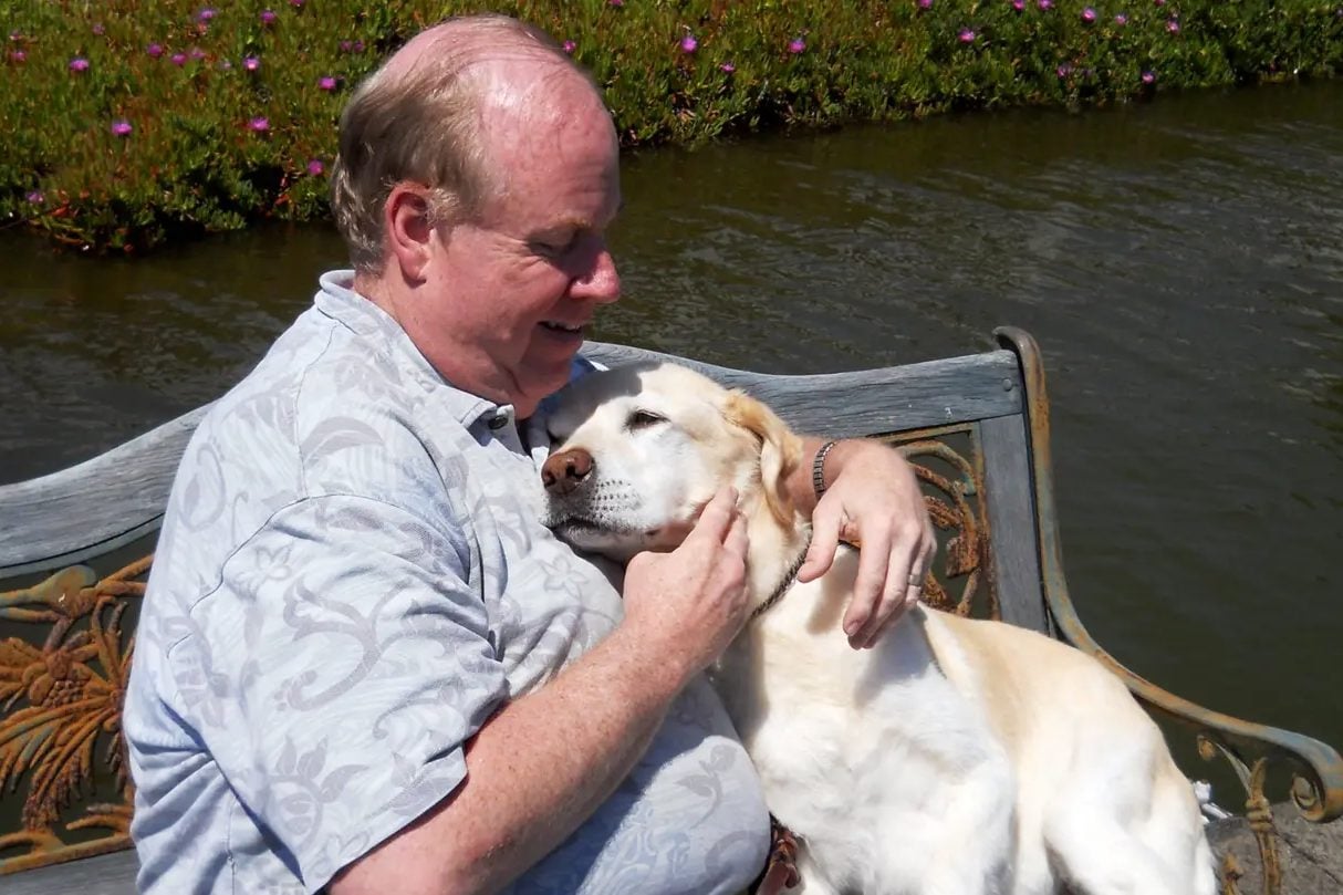 The image shows a man sitting on a bench with a large dog in his lap. The man is wearing a light blue shirt. There is a body of water in the background.