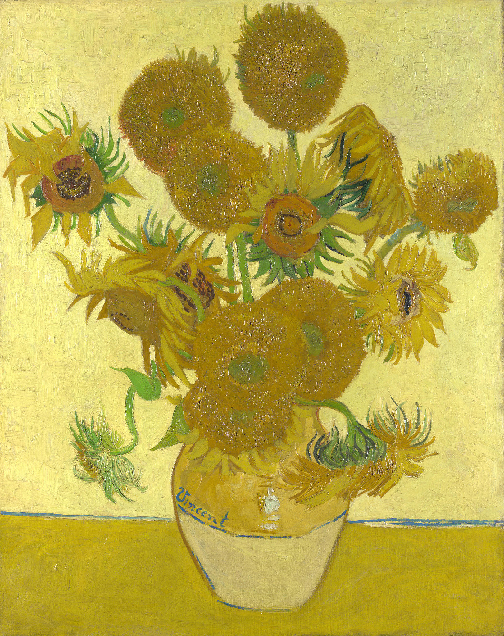 The image is a still life of a vase filled with sunflowers on a yellow table. The sunflowers are arranged in a circular pattern around the vase. The vase is made of a light-colored material and has a curved shape. The background of the image is a yellow wall.