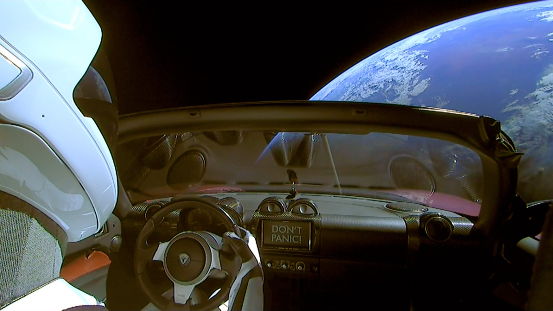A Tesla Roadster car with a dummy driver wearing a space suit floats in space with the Earth in the background.