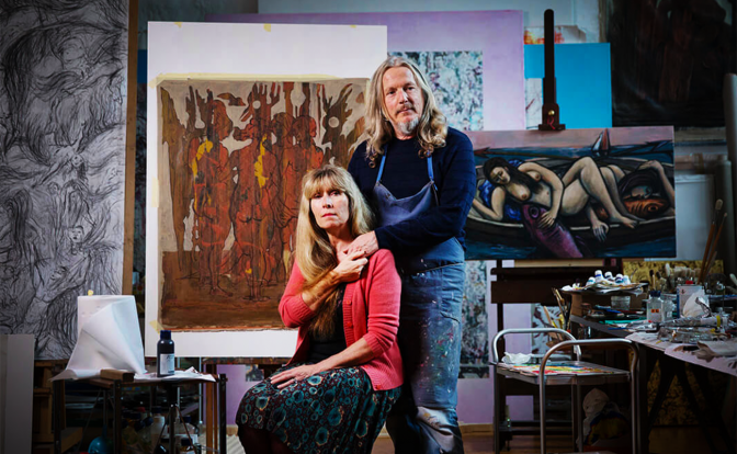 This photograph is of a man and a woman standing in front of an easel with a large painting on it. The man is wearing a blue shirt and jeans, while the woman is wearing a dark dress and a red sweater. They are both looking at the camera. There are paintings on the walls behind them. The room appears to be an artist's studio.