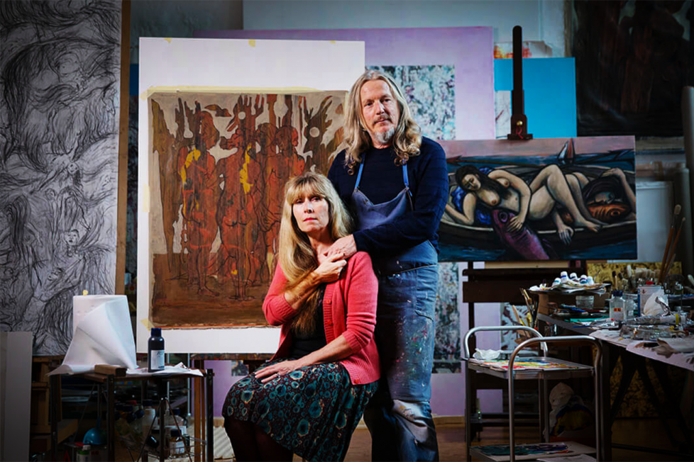 This photograph is of a man and a woman standing in front of an easel with a large painting on it. The man is wearing a blue shirt and jeans, while the woman is wearing a dark dress and a red sweater. They are both looking at the camera. There are paintings on the walls behind them. The room appears to be an artist's studio.