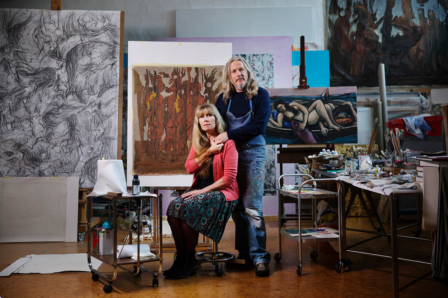 The image shows a man and a woman standing in front of an easel in an art studio. The man is wearing a blue shirt and the woman is wearing a black and red dress and a red sweater. They are both looking at the camera. There are several paintings on the walls behind them.