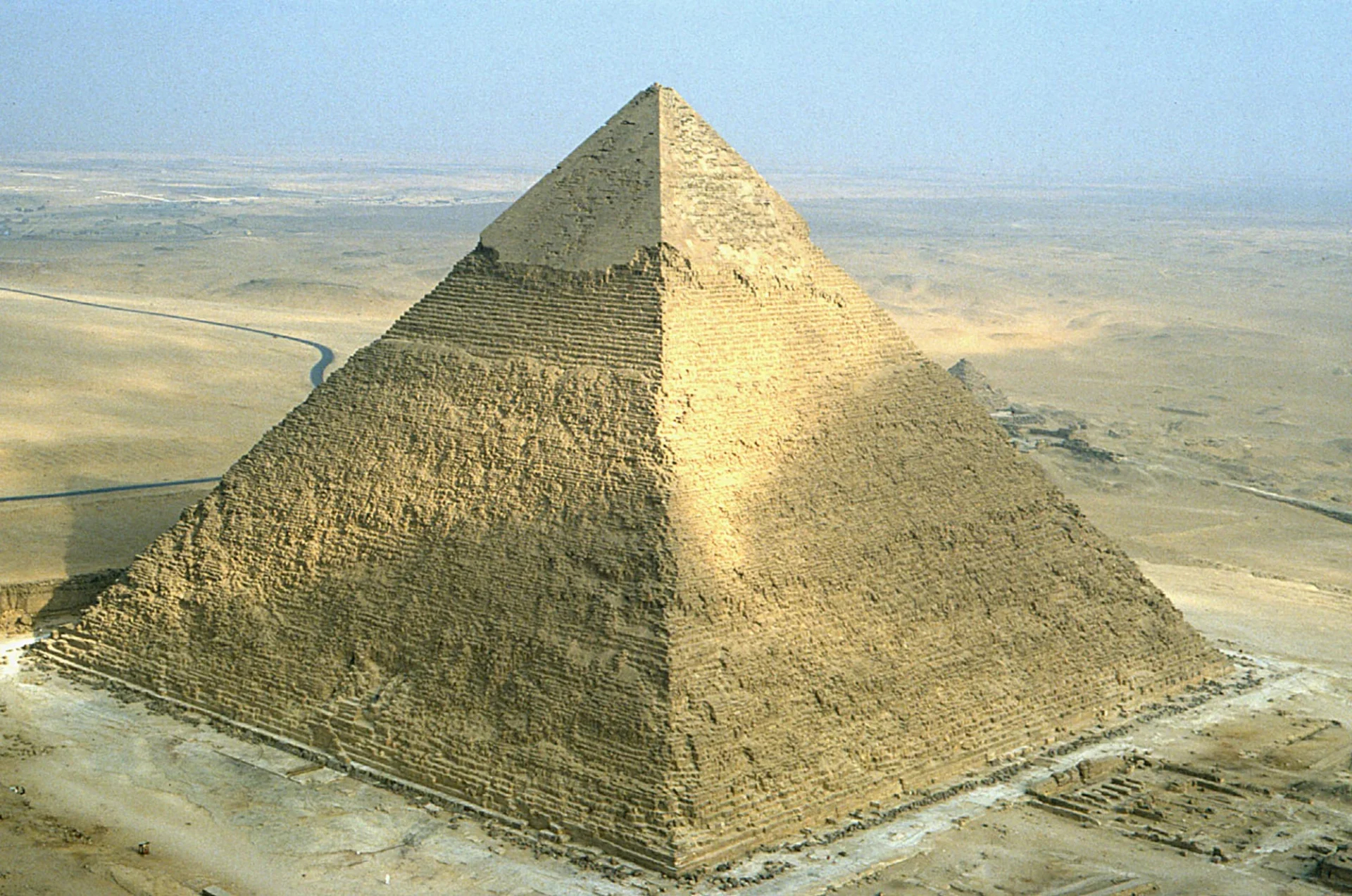 A photo of the Great Pyramid of Giza in Egypt, the oldest and largest of the three pyramids in the Giza pyramid complex, surrounded by a desert landscape and a hazy blue sky.
