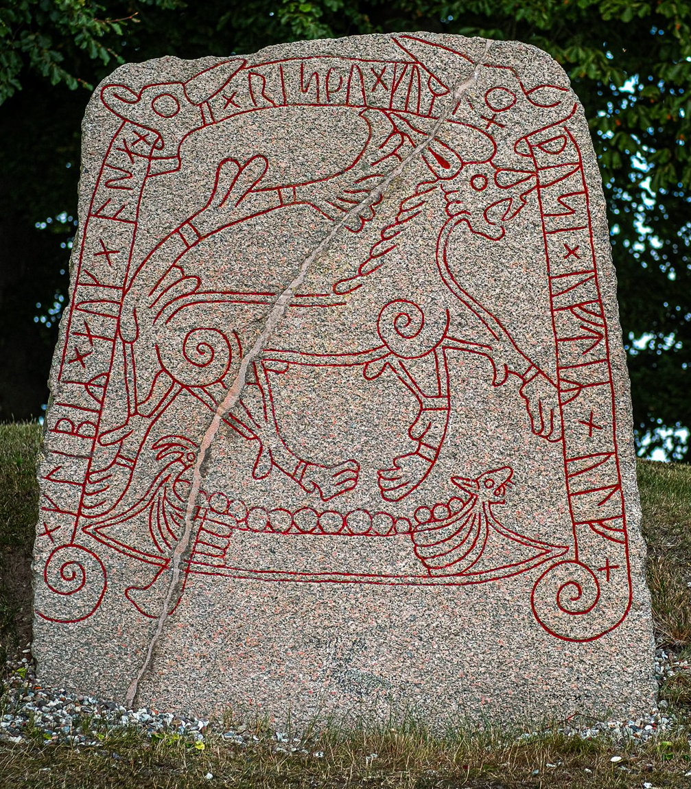 A photo of a large, granite runestone with intricate carvings on it. The runestone is standing upright on a grassy area with trees in the background. The carvings on the runestone are of a wolf, a ship, and various runes and symbols. The runes and symbols are carved around the edges of the stone. The stone appears to be weathered and aged.