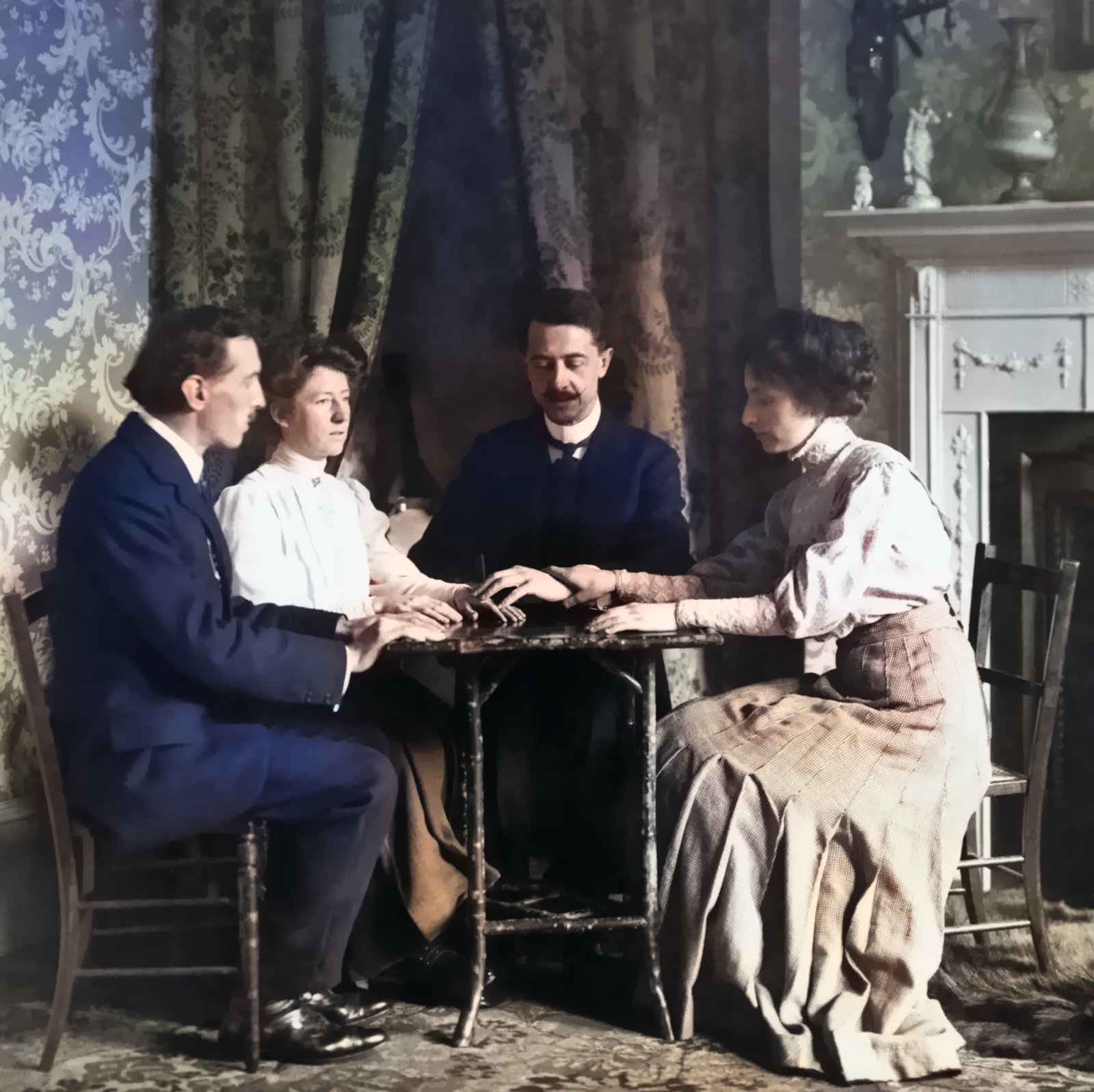 This is a black and white photo of four people sitting around a small table in a room with a fireplace and curtains. The people are dressed in formal Victorian clothing, with the two men wearing suits and the two women wearing dresses. The people are performing a séance led by Magician William Marriott.