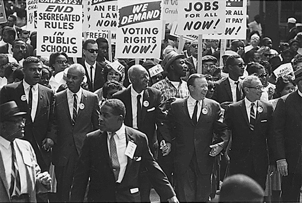 A black and white image of a protest march, showing the civil rights movement in the United States. The protesters are holding signs that read “End Segregated Public Schools”, “We Demand Voting Rights Now!”, “Jobs for All Now!” and “Freedom Now!”, expressing their demands for racial equality and justice.