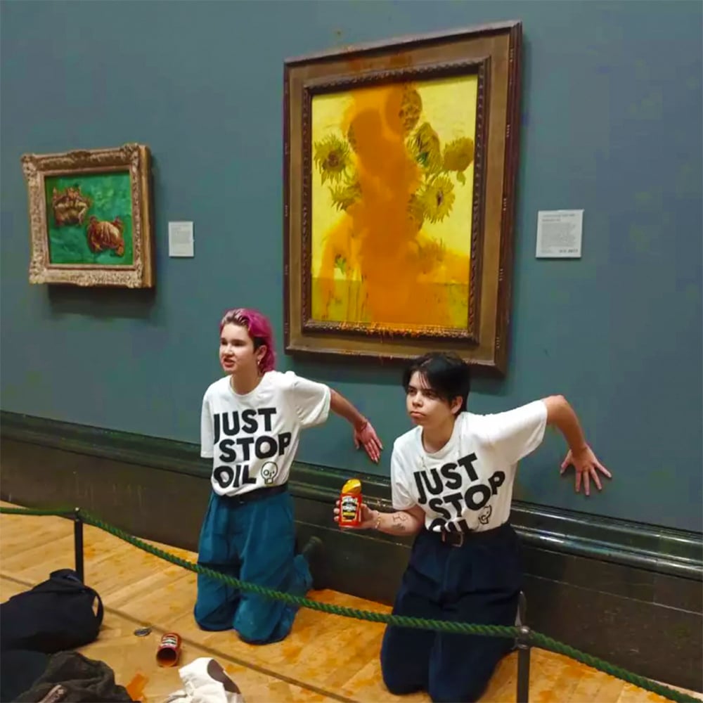 A photo of two people in a museum or gallery setting, protesting against oil consumption. The people are wearing white t-shirts with “Just Stop Oil” written on them, and are kneeling in front of a large, yellow, abstract painting with sunflowers on it. The painting is in a gold frame and is hanging on a green wall.