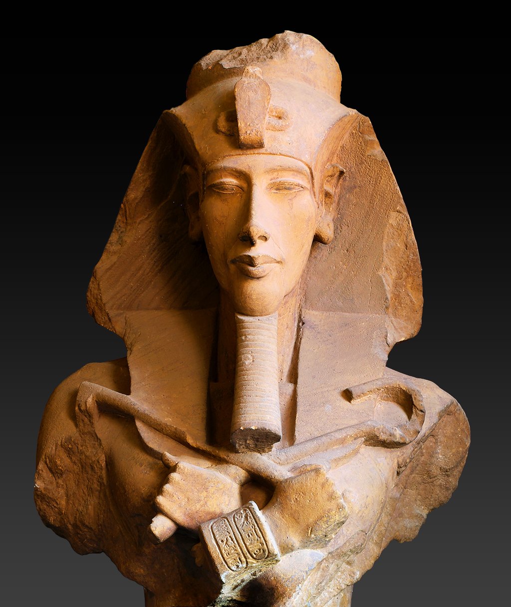 A stone sculpture of a Amenhotep IV/Akhenaten bust. His hands are crossed at the chest. The sculpture is on a black background.