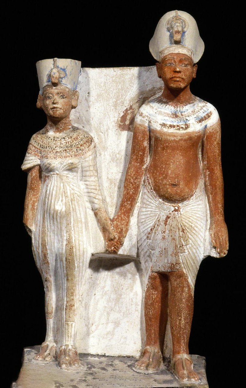 Statuette of Akhenaten and Nefertiti: king (standing, wearing a pleated skirt with an apron front, blue crown, uraeus, broad collar necklace, sandals, holding hand) and queen (standing, in pleated dress, Amarna crown, broad collar necklace, earrings, sandals).