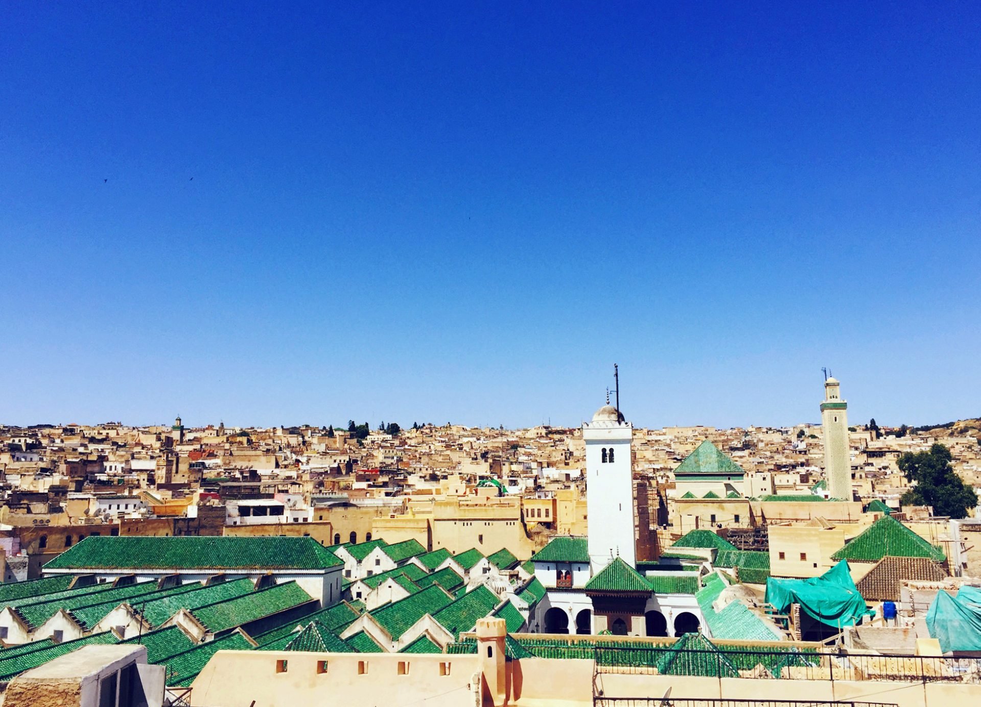 A view of the historic city of Fez in Morocco, with the white and beige buildings and green rooftops of the Al-Qarawiyyin Mosque and school complex in the center. The mosque has two tall minarets, one with a clock tower and the other with a green dome. The sky is clear and blue.