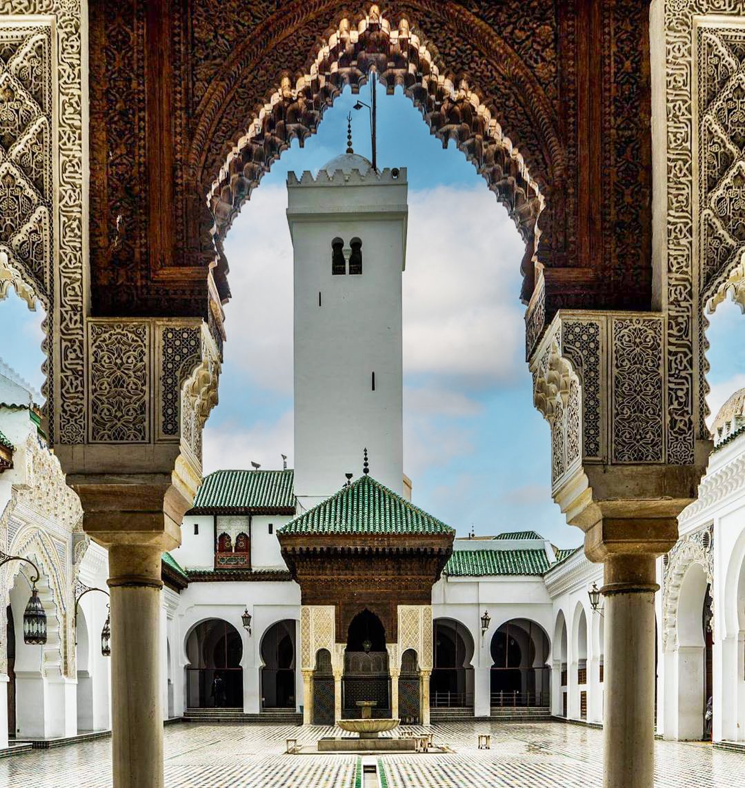 Al-Qarawiyyin courtyard with green and white floor mosaics, a fountain, arches, and a view of a mosque and its minaret in the background.