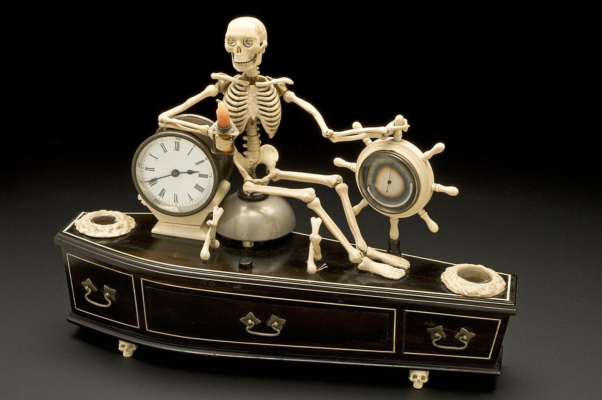 An English “memento mori” alarm clock shaped like a coffin. On top lies an ivory skeleton, one hand holding a sand timer and a candle, the other on a ship's wheel containing a thermometer and barometer. The coffin rests on four ivory skulls and has two candle wells. When the alarm sounds, the skeleton's jaw opens and eyes roll, emphasizing reminders of life's brevity and impending death.