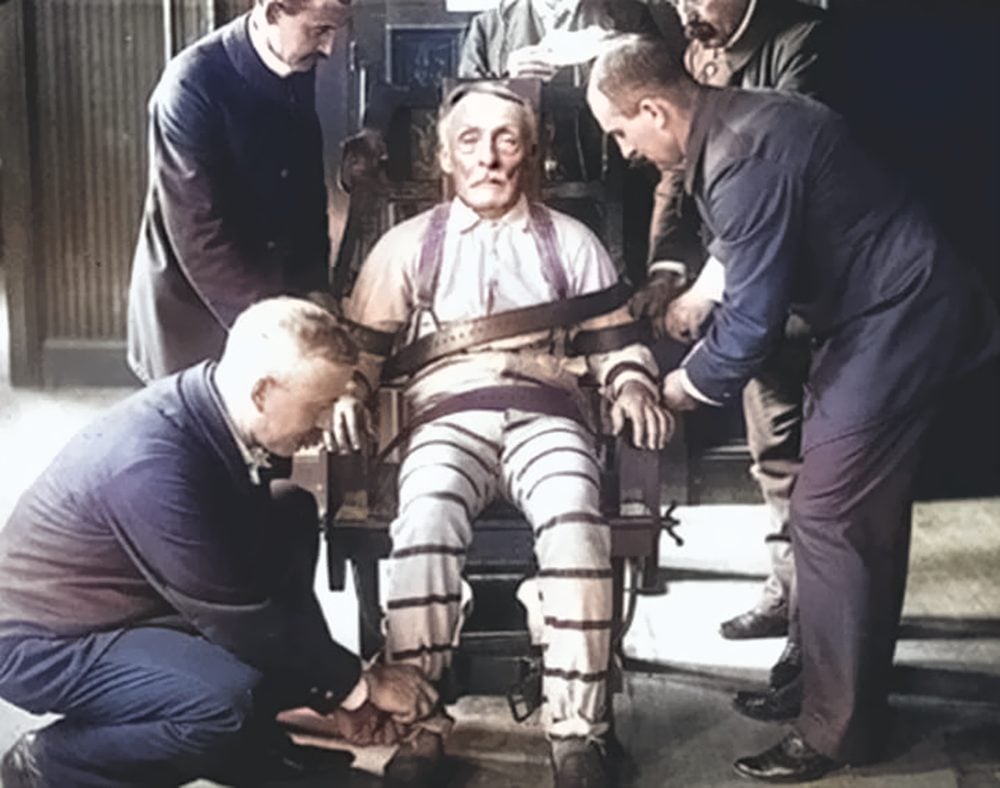 A photograph of an elderly man in a white prison uniform sitting in an electric chair. His eyes are closed. He is being prepared for execution by four prison guards.